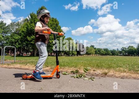 Un jeune garçon joue avec son scooter et le pousse lors d'une journée ensoleillée dans le parc. Ciel bleu et arbres verts. Enfant énergique et actif. Banque D'Images