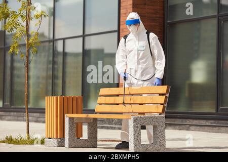 Portrait complet de l'employé portant une combinaison de protection pulvérisant des produits chimiques sur un banc à l'extérieur pendant la désinfection ou le nettoyage, espace de copie Banque D'Images