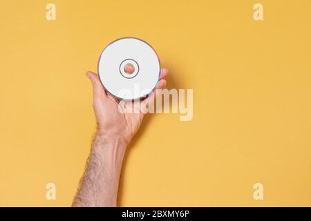 Main de sexe masculin tenant un CD-DVD devant un fond jaune, modèle de maquette modifiable pour votre conception, chemin de sélection d'étiquette inclus Banque D'Images