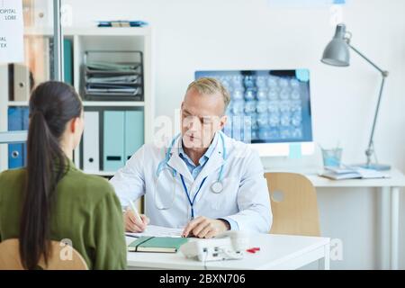 Portrait d'un médecin de sexe masculin mature écrit sur un presse-papiers tout en étant assis au bureau et en consultant une patiente dans une clinique moderne, espace de copie Banque D'Images