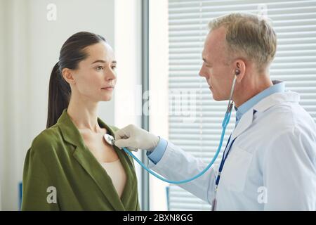 Portrait de vue latérale d'un médecin mature écoutant le souffle et le battement de coeur d'une jeune femme via le stéthoscope tout en examinant le patient en clinique Banque D'Images