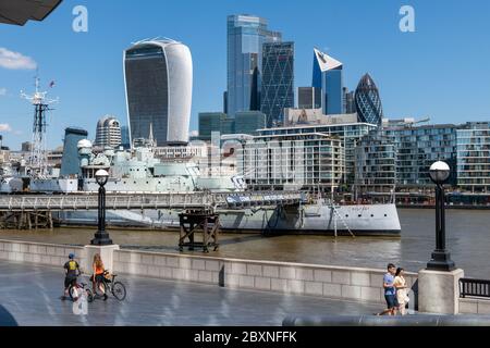 Visiteurs de la rive sud en profitant d'une belle journée chaude au bord de la Tamise avec vue sur le HMS Belfast et la ville de Londres. Angleterre.Royaume-Uni. Banque D'Images