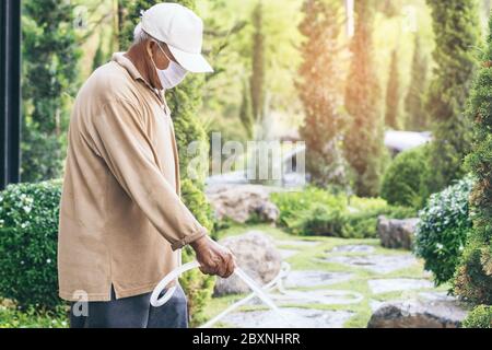 Un homme âgé en quarantaine porte un masque facial pour empêcher la propagation du virus Corona (Covid-19) arroser des plantes pour faire de l'exercice dans le jardin à la maison. Ne Banque D'Images