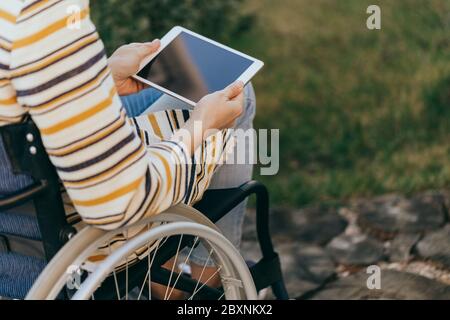 Femme habillée de façon tendance dans un fauteuil roulant avec une tablette moderne dans la main marche dans la rue. Personne méconnaissable. Banque D'Images