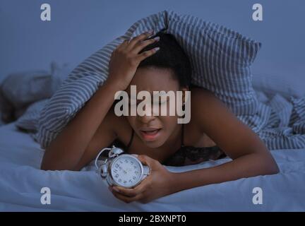 Nuit sans sommeil. Triste afro-américaine fille appuie main à la tête et tenant le réveil Banque D'Images