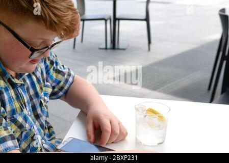 Garçon aux cheveux rouges avec téléphone portable buvant dans un verre d'eau glacée au citron. Enfant avec des lunettes vêtues d'une chemise à carreaux regarde le smartphone et d Banque D'Images