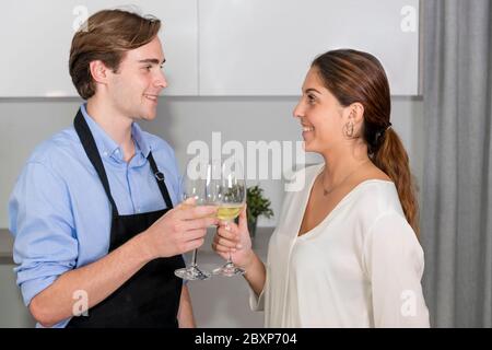 Un jeune couple se souriant et se faisant griller avec deux verres de vin blanc dans la cuisine sur un fond délabsé. Cuisiner à la maison et s'amuser c Banque D'Images