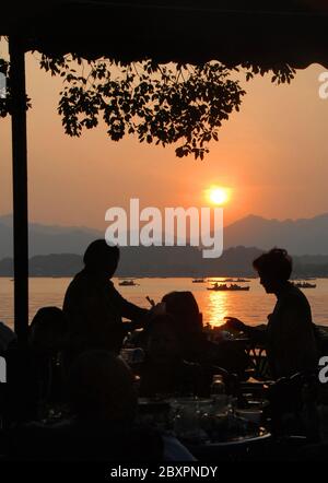 Coucher de soleil sur le lac de l'Ouest (Xi Hu) à Hangzhou, province du Zhejiang, Chine. Vue sur le lac de l'Ouest, Hangzhou avec silhouettes de personnes assises dans un café. Banque D'Images