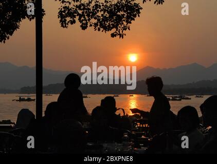 Coucher de soleil sur le lac de l'Ouest (Xi Hu) à Hangzhou, province du Zhejiang, Chine. Vue sur le lac de l'Ouest, Hangzhou avec silhouettes de personnes assises dans un café. Banque D'Images