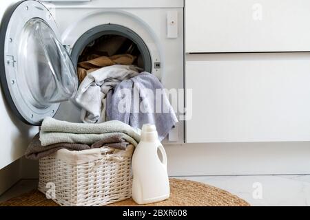 Chargement d'un lave-linge et lavage de vêtements. Un panier à linge et un gel à linge devant un lave-linge intégré dans la cuisine. Banque D'Images