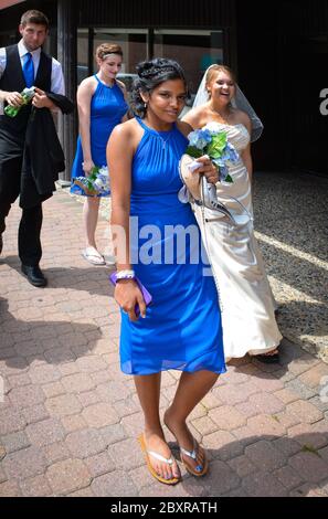 Une demoiselle d'honneur en robe bleue porte des talons hauts et des fleurs pour rire jeune femme en robe blanche et voile de mariée, sur le trottoir à St. Cloud, MN, USA Banque D'Images