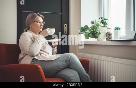 Femme caucasienne âgée avec lunettes assise dans un fauteuil et buvant un café tout en portant des lunettes et en regardant une leçon sur un ordinateur portable Banque D'Images