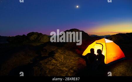 Jeune couple assis près d'une tente illuminée et regardant l'un l'autre à Beautirul Evening dans les montagnes. Concept aventure et voyage en famille. Banque D'Images