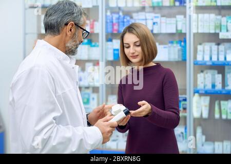 Pharmacien offrant des produits médicaux pour les femmes clients en pharmacie. Belle femme tenant des bouteilles de cosmétiques blanches, choix. Vue arrière d'un chimiste méconnaissable portant des lunettes et un blouse de laboratoire blanche. Banque D'Images