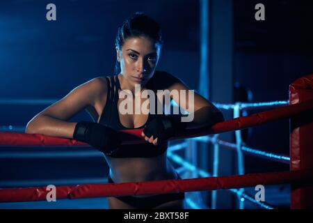 kickboxer femme professionnelle sérieuse jeune portant des vêtements de sport et des bandages noirs sur les mains, posant dans une atmosphère sombre. Chasseur ayant le repos après l'entraînement, se pencher sur la corde et regarder la caméra. Banque D'Images