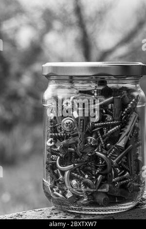 Photo noir et blanc d'un pot en verre de boulons et écrous assortis, en gros plan sur du verre flou sur fond bokeh avec espace de copie. Métal aussi Banque D'Images