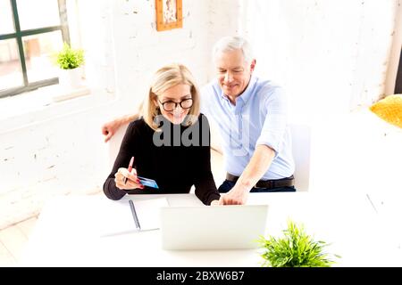 Photo en grand angle d'un couple senior heureux qui fait des finances à la maison. Femme âgée attrayante tenant une carte bancaire dans sa main tandis que l'homme senior assis derrière le Banque D'Images