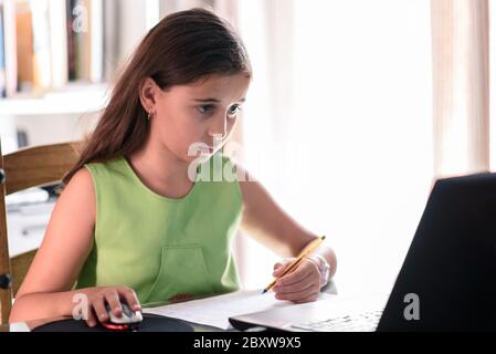 Une jeune fille concentrée fait ses devoirs à la maison devant un ordinateur portable. Éducation, apprentissage et concept scolaire. Lumière naturelle Banque D'Images
