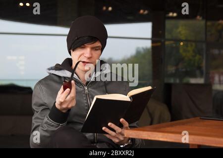 Vue arrière sur un jeune homme à cheveux noirs qui pierrait en repiquant un livre et en fumant le tabacco pipe dans un café extérieur Banque D'Images