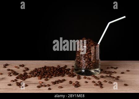 Verre à whisky plein de grains de café rôtis avec paille sur un panneau de bois sur fond sombre Banque D'Images