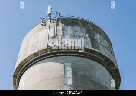 Tour d'eau en béton coulé très abîmé, vu contre un ciel bleu clair. Les mâts de télécommunication 5G récemment installés sont visibles au sommet de la tour. Banque D'Images