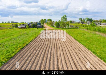 Labourer des sillons de terre pour planter des plantes agronomiques dans la campagne loin d'un village résidentiel sur une colline, vue aérienne d'en haut Banque D'Images