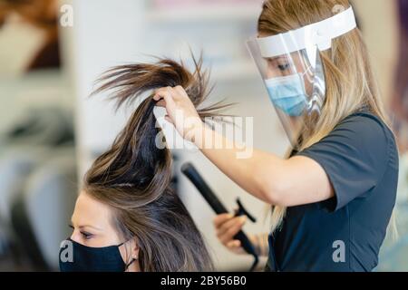 Coiffeur, protégé par un masque, faisant des vagues dans les cheveux de son client avec un fer à repasser dans un salon. Concepts d'affaires et de beauté Banque D'Images