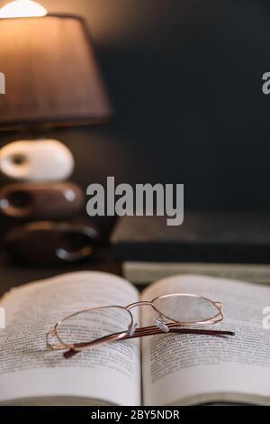 La vie est encore plus longue avec les vieux livres et les lunettes de lecture éclairées par la lumière de nuit. Ambiance calme le soir dans une chambre marron foncé. Banque D'Images