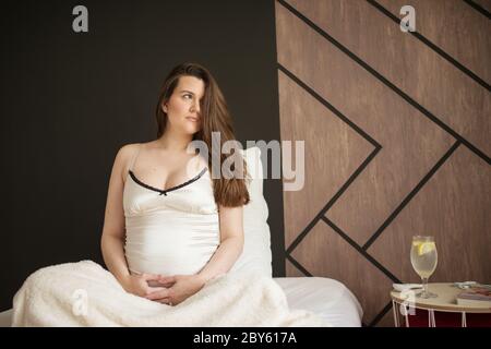 Grossesse, repos, personnes et concept d'attente. Une femme enceinte heureuse assise sur le lit et touchant son ventre à la maison. Banque D'Images