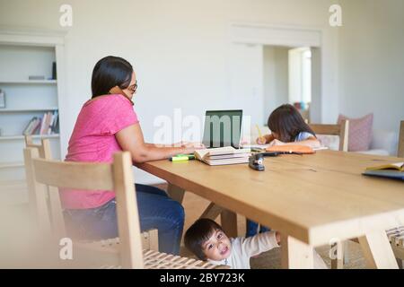 Mère travaillant sur un ordinateur portable avec des enfants jouant et faisant leurs devoirs Banque D'Images