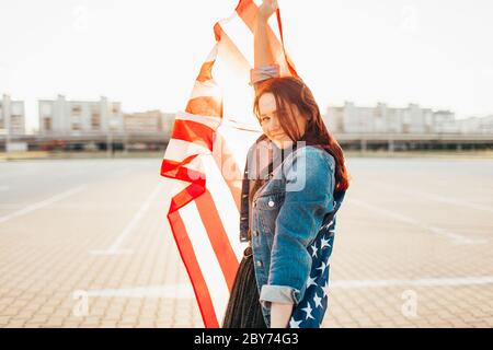 Jeune femme à poil rouge enveloppée de drapeau national des états-unis au soleil. Rétroéclairage doux. Jour de l'indépendance, rêve américain, concept de liberté Banque D'Images