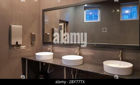Robinets chromés avec lavabos ronds blancs dans une toilette publique avec un grand miroir et des murs gris, intérieur moderne d'une toilette publique Banque D'Images