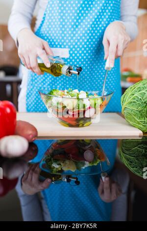 Gros plan de la femme en tablier préparant une salade de légumes et ajoutant de l'huile d'olive au bureau de cuisine. Banque D'Images