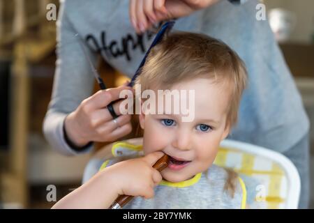 Gros plan jeune adulte de race blanche mère faire la coupe de cheveux fot mignon enfant adorable fils adorable garçon à la maison en raison de la quarantaine et de verrouillage. Maman coupant les cheveux de l'enfant Banque D'Images