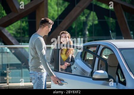 Jeune jolie femme ouvrant la porte de la voiture, parlant au jeune homme et souriant Banque D'Images