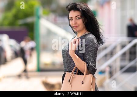 Élégante femme aux cheveux sombres qui marche à l'extérieur de la ville avec un sac à main en cuir sur son bras tournant pour regarder l'appareil photo avec un sourire amical et silencieux Banque D'Images
