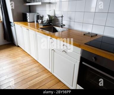 Appareils et meubles: Armoires blanc lumineux avec comptoir en bois, cuisinière électrique, plaque à induction, robinet, évier et porte-plats contre le bois dur Banque D'Images