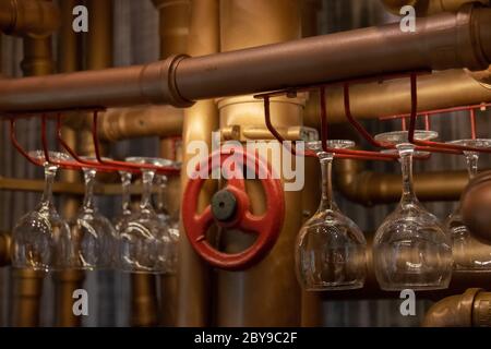 Les lunettes de vin vides transparentes brillantes sont suspendues à l'envers sur les tablettes de bar en tuyaux d'eau peints en bronze avec une vanne ronde rouge au centre Banque D'Images