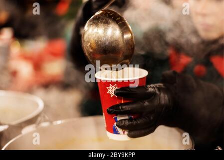 Un vendeur de rue méconnu verse du vin chaud chaud dans une tasse jetable. Boisson traditionnelle d'hiver par temps froid. Salon de Noël. Gros plan Banque D'Images