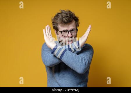 Un hipster insatisfait dans un ancien pull montre un mouvement fermé chemin avec les bras croisés. Studio, fond jaune. Banque D'Images