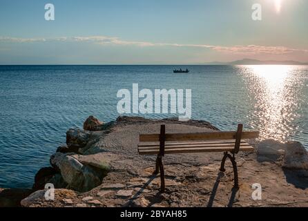 Coucher de soleil à Afissos, Grèce, avec un banc vide en premier plan et mer Égée et un bateau de pêche avec une silhouette de pêcheur en arrière-plan Banque D'Images