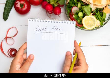 Vue de dessus des mains de femme tenant bloc-notes avec des mots manuscrits régime Plan au-dessus de la table blanche avec légumes et salade Banque D'Images