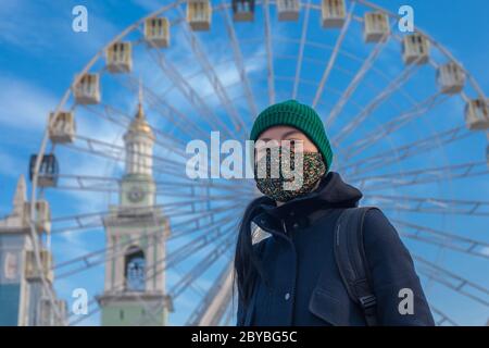 Kiev, Ukraine - 26 mars 2020: La roue de Ferris sur fond de ciel bleu et une fille dans un masque protecteur sur son visage, sur la place Kontraktova sur Podil dans le centre de Kiev. Concept de quarantaine Banque D'Images