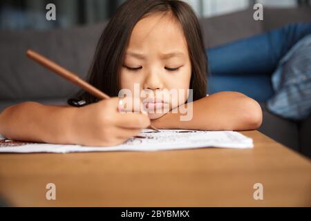 Photo gros plan de petite fille asiatique concentrée avec de longs cheveux dessinant tout en étant assis à la table à la maison Banque D'Images