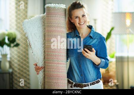 bonne femme de ménage d'âge moyen dans une chemise de jeans et un pantalon blanc avec moquette blanche à la recherche d'un service de nettoyage de tapis sur un smartphone dans la vie moderne Banque D'Images