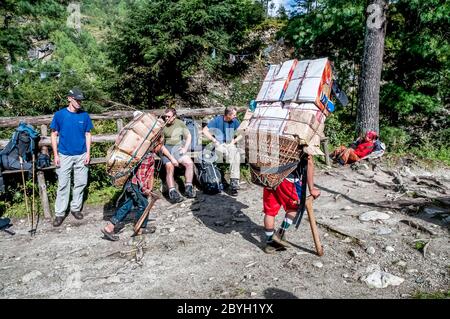 Népal. Island Peak Trek. Un porteur lourdement chargé passe par un groupe de randonneurs européens en train de faire une pause en route vers la ville commerciale de Namche Bazar Banque D'Images