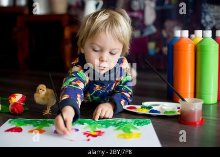 Doux tout-petit blond enfant, garçon, peinture avec des couleurs, petits poussins qui marchent autour de lui, faisant des imprimés amusants sur son papier Banque D'Images