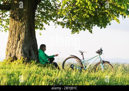 Une fille solitaire est assise sous un grand vieux arbre près d'un vélo. Paysage d'été vert en contreface de la ville Banque D'Images