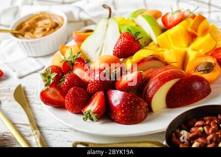 Plateau de fruits colorés de vacances de fraises, de mangue, de pommes, de poires, d'abricots, avec beurre d'arachide et d'arachides sur une assiette blanche sur un dos en bois blanc Banque D'Images