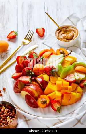 Assiette de fruits snack de fruits frais et de baies : fraises, mangue, pommes, poires, abricots, beurre d'arachide et arachides sur une assiette blanche sur une wh Banque D'Images